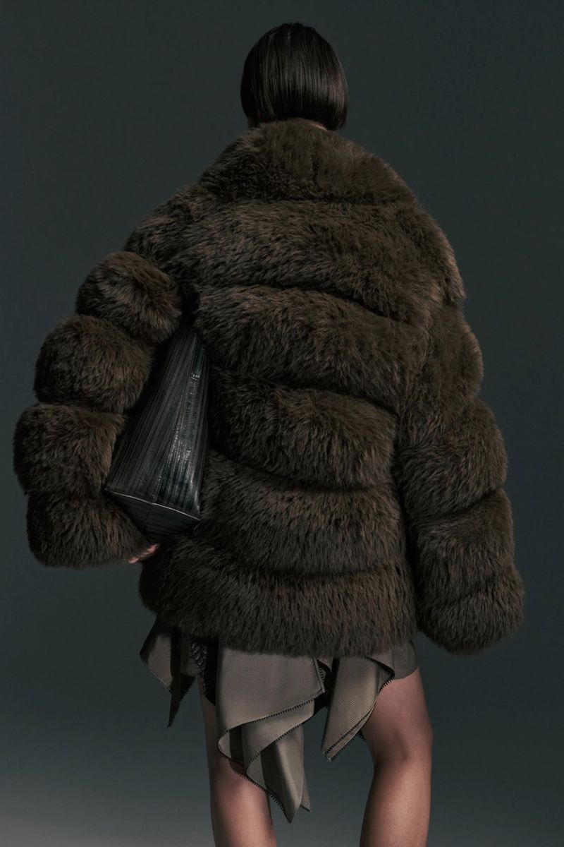 Rear view of a model in an oversized dark brown fur jacket with a black tote bag tucked underarm against a dark grey studio background.