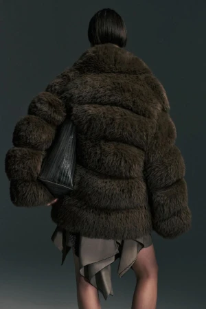 Rear view of a model in an oversized dark brown fur jacket with a black tote bag tucked underarm against a dark grey studio background.