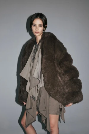 Model wearing an oversized dark brown fur jacket and draped taupe dress against a grey backdrop.