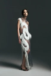 Model wearing a sheer lace dress with white curved fringe detailing and black strappy heels against a dark grey studio background.