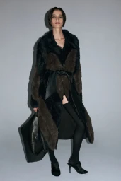 Model wearing a belted two-tone faux-fur coat, black tights, and heels while holding a large black bag against a grey background.