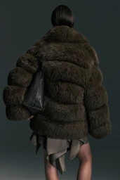 Rear view of a model in an oversized dark brown fur jacket with a black tote bag tucked underarm against a dark grey studio background.