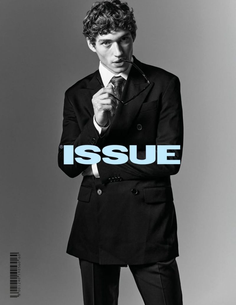 Portrait of a young man in a black suit holding glasses in front of an 'Issue' magazine cover in a neutral studio backdrop.