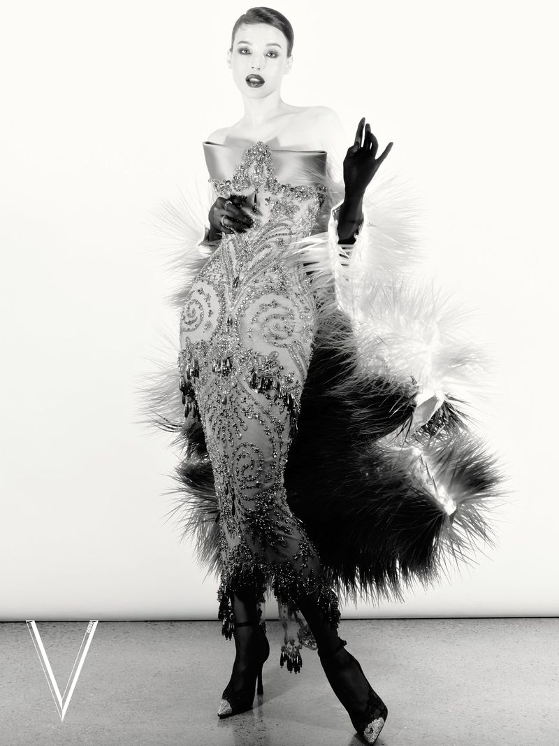 Monochrome fashion portrait of a woman in an ornate beaded gown, opera gloves, and feathered stole against a white seamless backdrop.