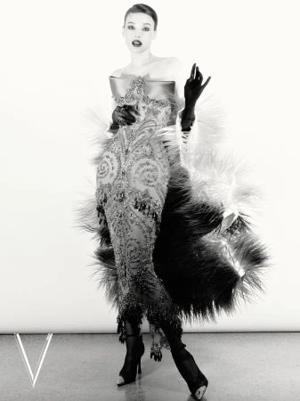 Monochrome fashion portrait of a woman in an ornate beaded gown, opera gloves, and feathered stole against a white seamless backdrop.