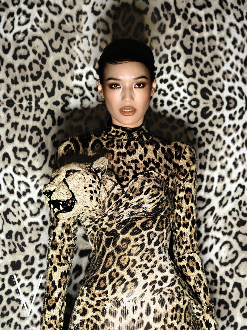 Model wearing a high-neck leopard print dress with a sculptural feline head against a matching animal print backdrop.