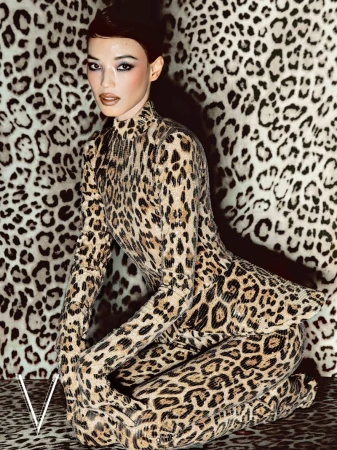 An East Asian woman kneeling in a full-body leopard print outfit against a matching leopard print patterned backdrop.