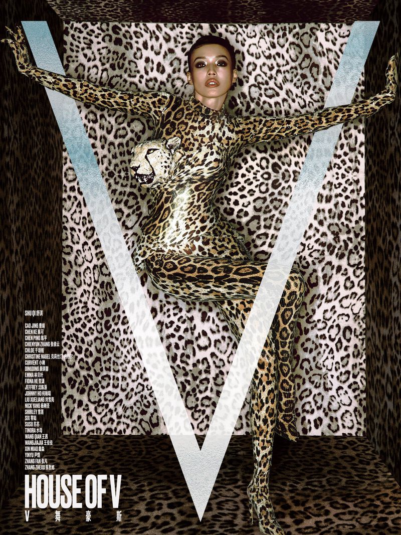 Shu Qi posing in a full leopard-print catsuit with a sculpted cheetah head against a matching leopard-patterned enclosure behind a large V logo.