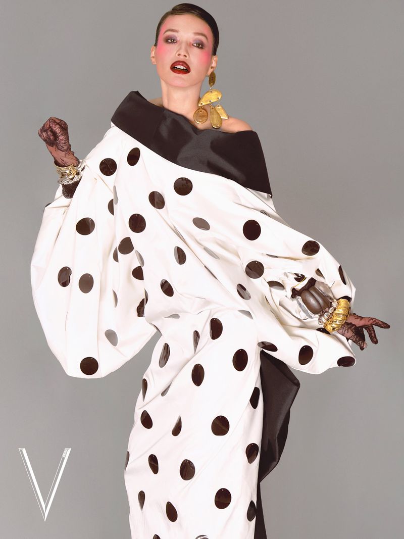 A woman poses in a black and white polka dot dress with gold jewelry, against a grey background, displaying a high-fashion editorial style.