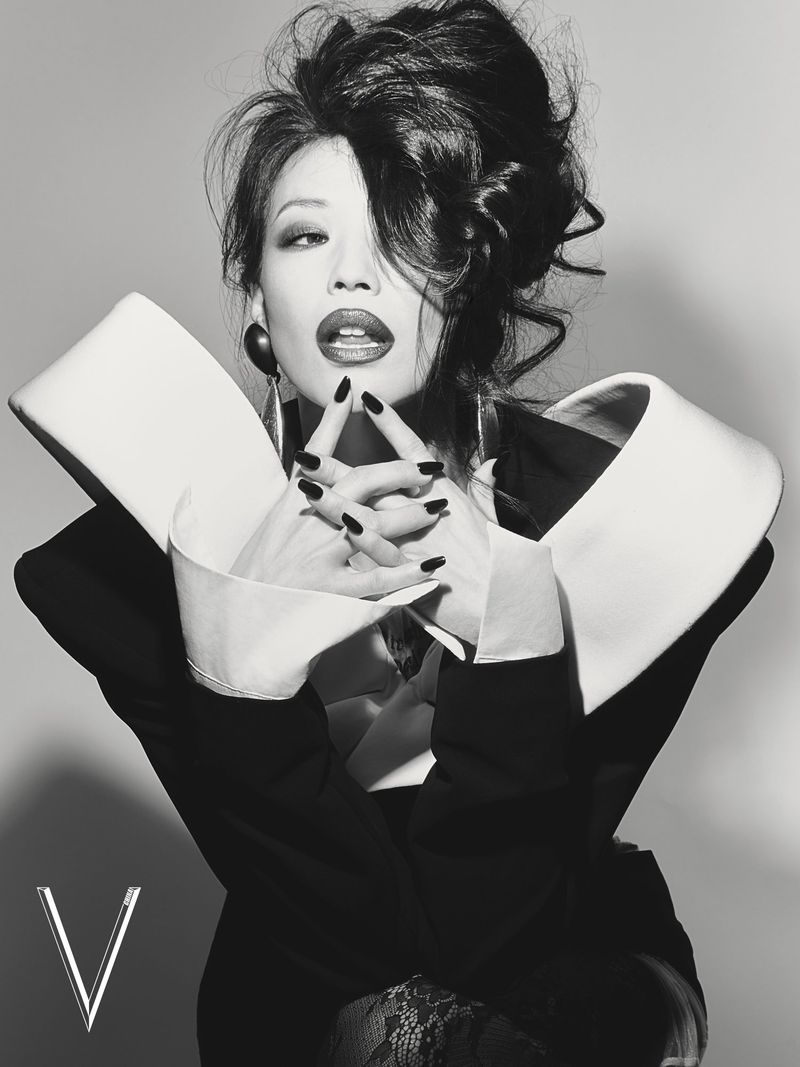 Model with a dramatic voluminous updo posing in a black blazer with an oversized sculptural white collar against a grey studio backdrop.