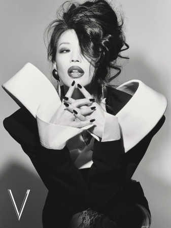Model with a dramatic voluminous updo posing in a black blazer with an oversized sculptural white collar against a grey studio backdrop.