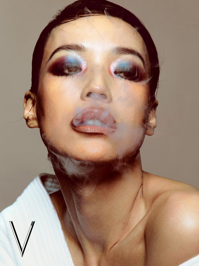 East Asian model with bold colorful eyeshadow and slicked-back hair exhaling smoke in a white robe against a studio backdrop.