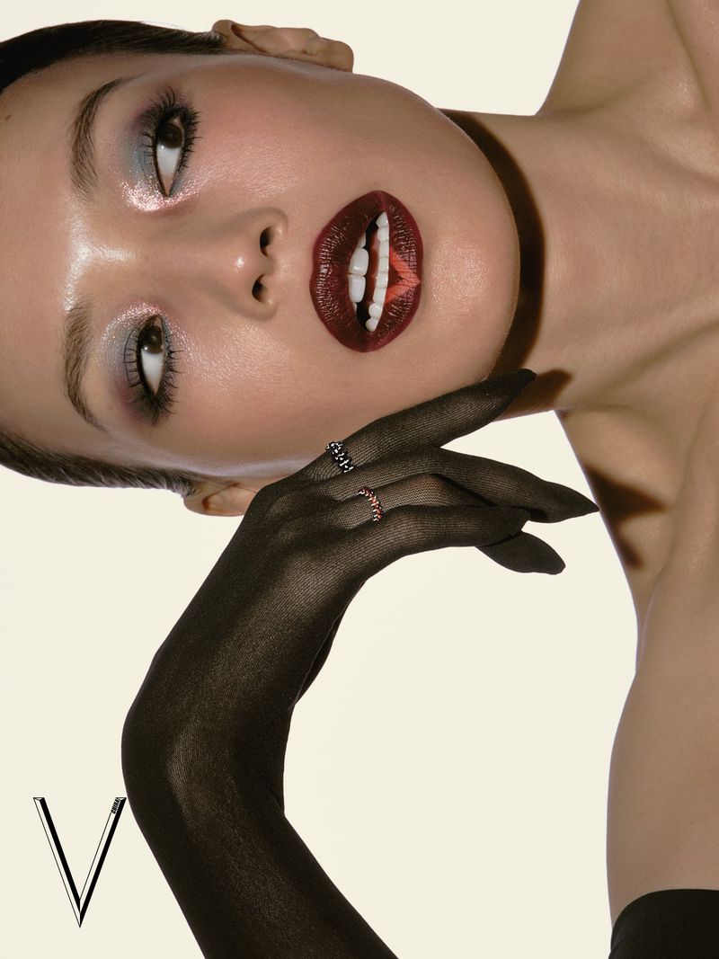 Close-up beauty portrait of a woman with dramatic dark red lips wearing sheer black opera gloves against a cream studio backdrop.