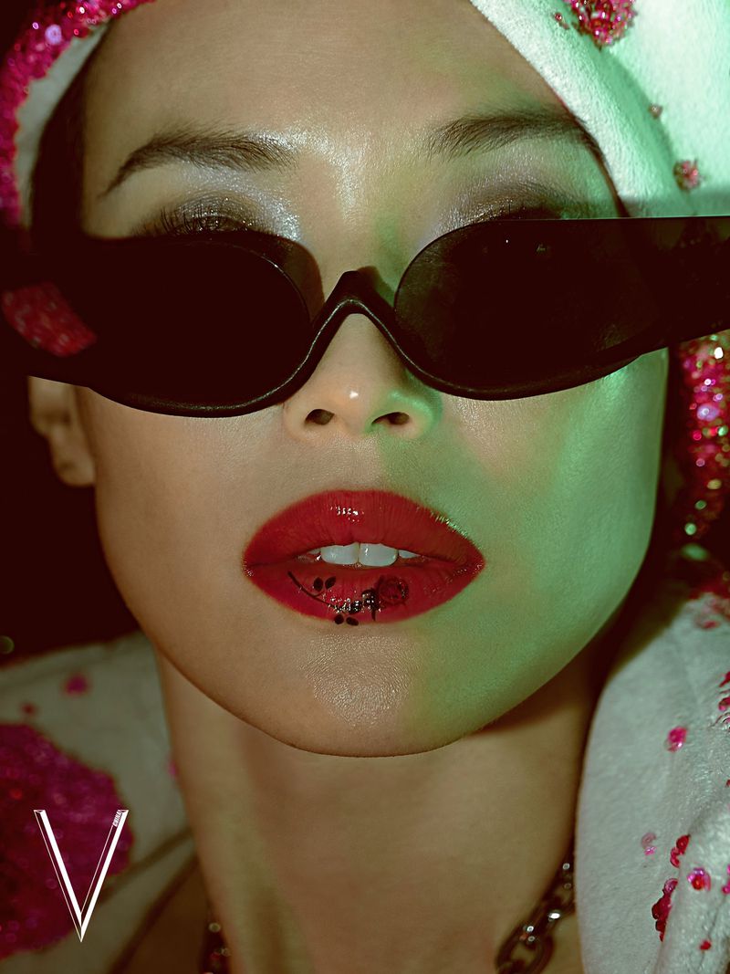Close-up portrait of an East Asian woman wearing black cat-eye sunglasses, bold red lipstick with floral art, and a white sequined hood.
