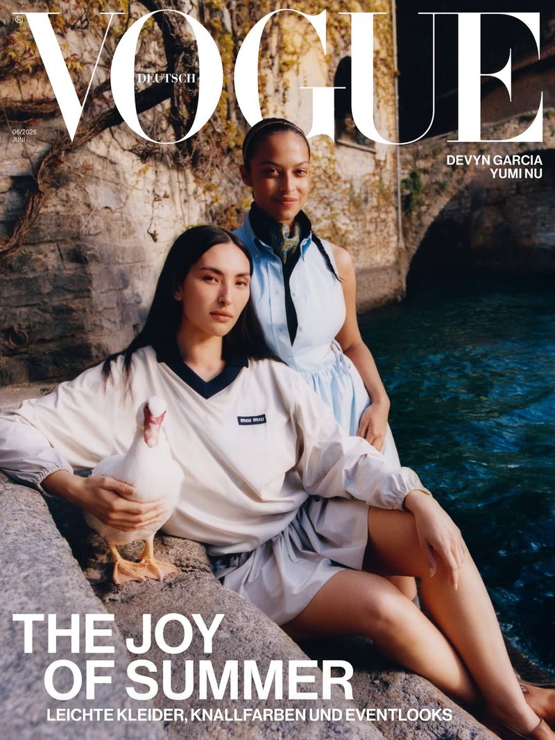 Two women pose on rocks by water for a Vogue Deutsch cover; one holds a white duck in an outdoor setting with stone architecture.