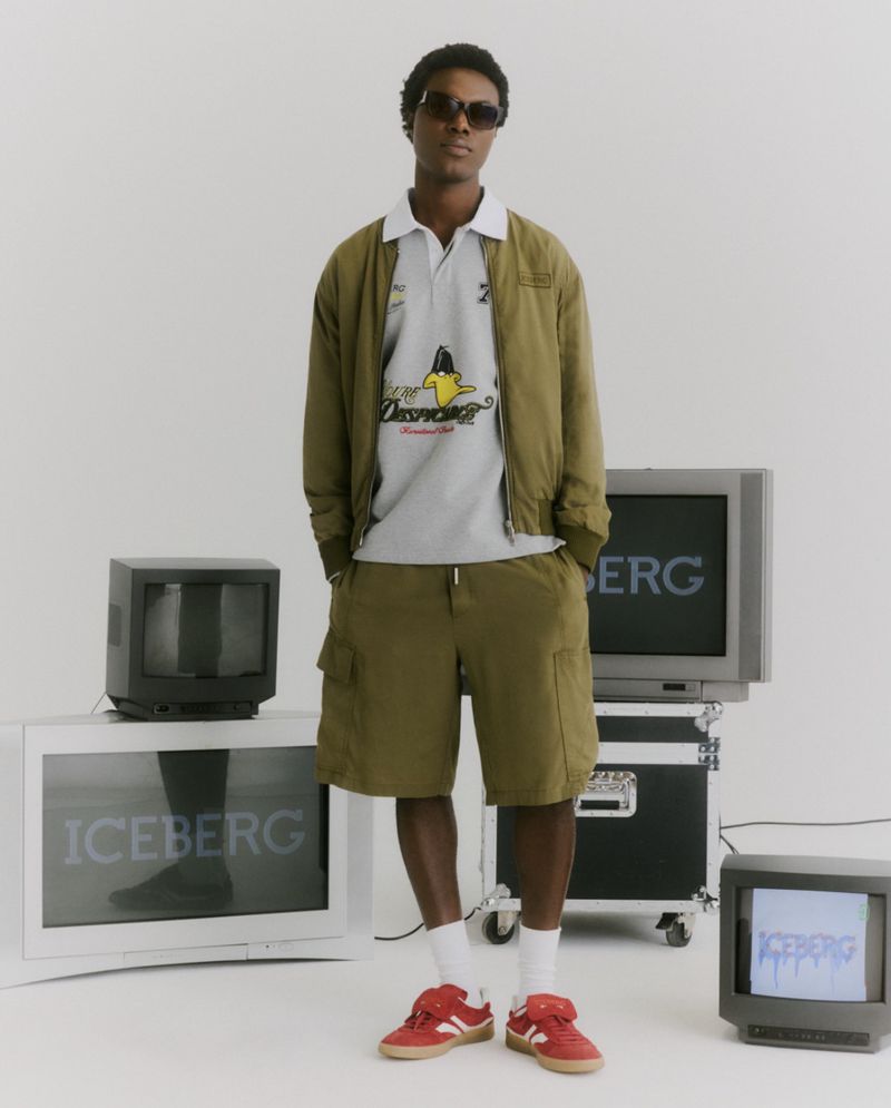 Model Mensah Benjamin standing in an olive green jacket, cargo shorts, and red sneakers surrounded by CRT television monitors.
