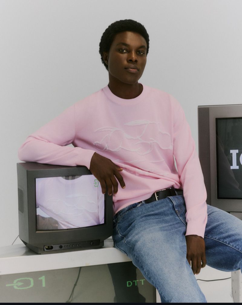 Model Mensah Benjamin wearing a pastel pink sweater and blue jeans leaning on vintage CRT televisions in a studio setting.