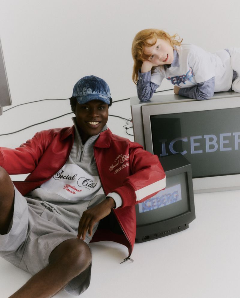 A Black male model in a red jacket and blue cap sits beside vintage CRT monitors displaying the Iceberg logo on a white seamless.