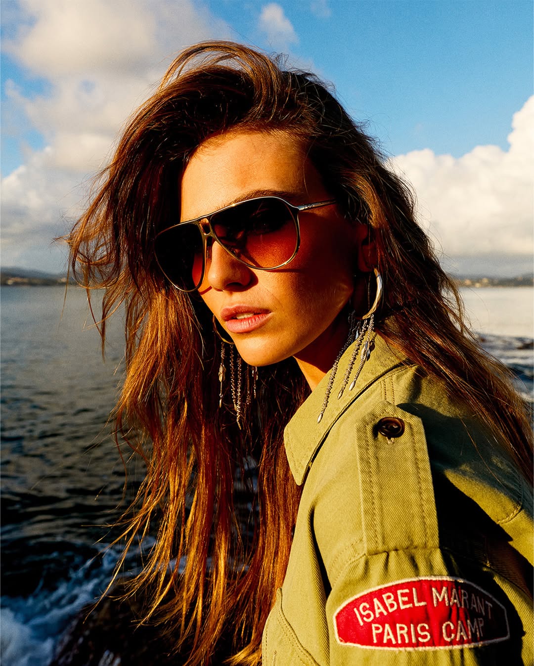 Model Mia Armstrong wearing tinted aviator sunglasses, tassel earrings, and an olive utility jacket by the ocean.