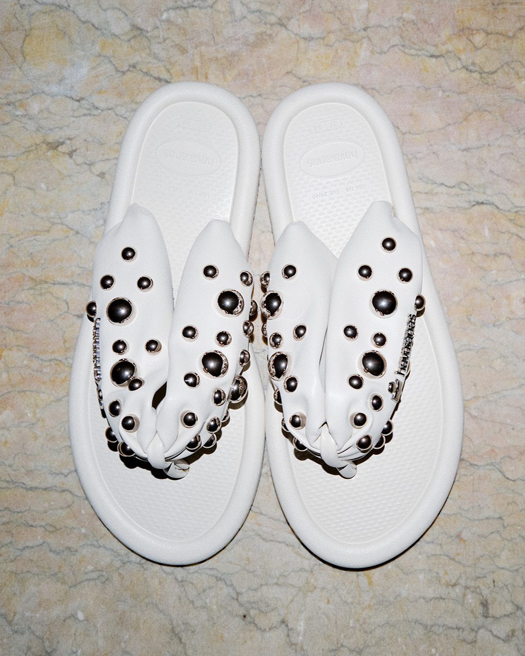 Pair of white Isabel Marant x Havaianas flip-flop sandals with gathered studded straps resting flat on a polished marble tile floor.