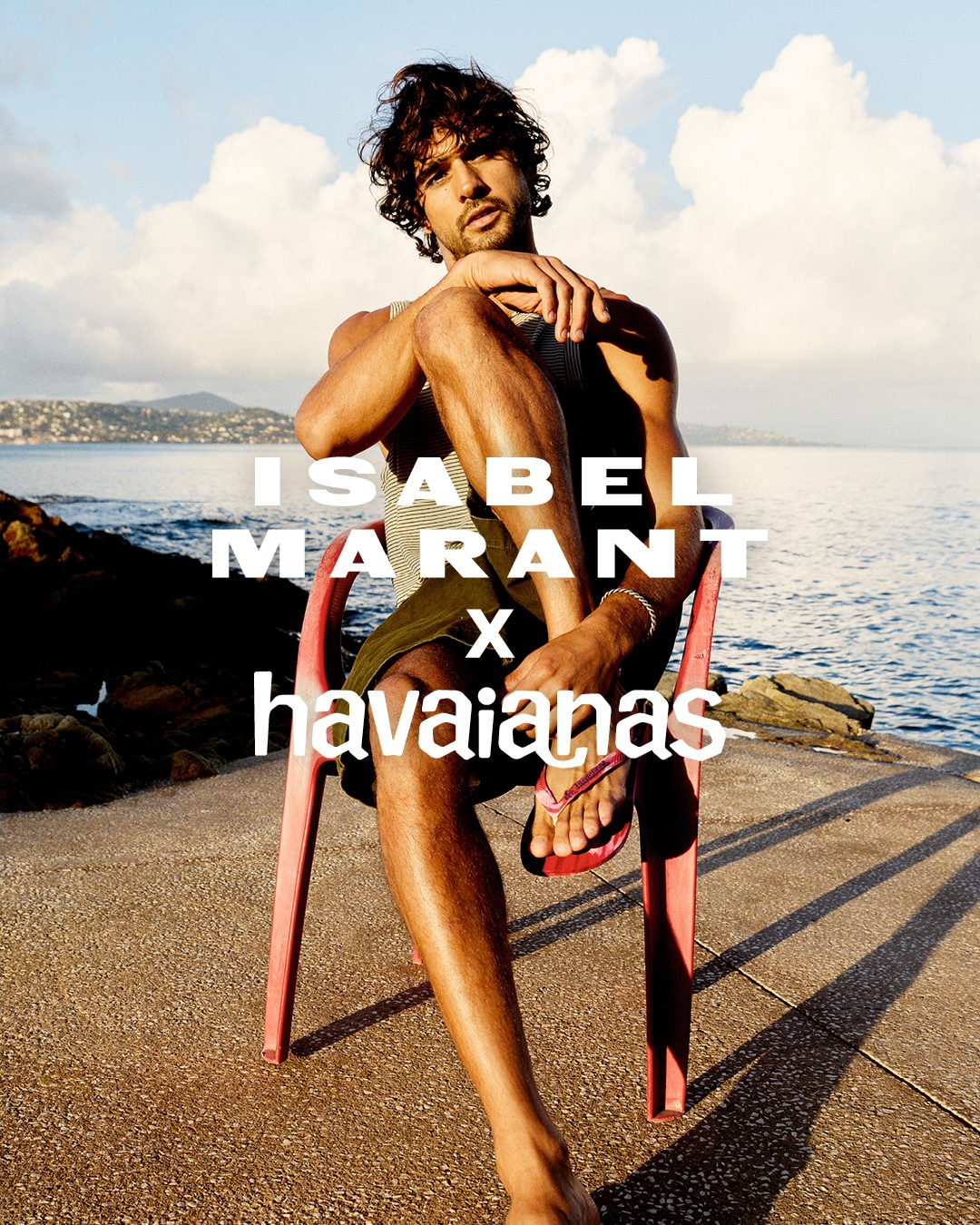 Model Marlon Teixeira seated on a red plastic chair by the coast for the Isabel Marant x Havaianas campaign.