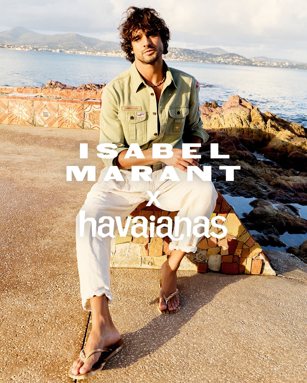 Marlon Teixeira wearing an olive green button-up shirt, cream trousers, and flip-flops seated on a stone ledge by the coast.