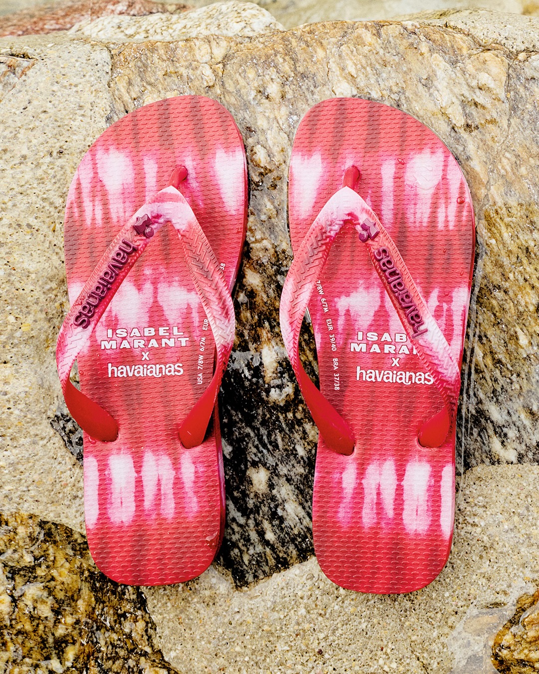 A pair of red and white tie-dye Isabel Marant x Havaianas flip-flops resting vertically on a textured wet rock surface.