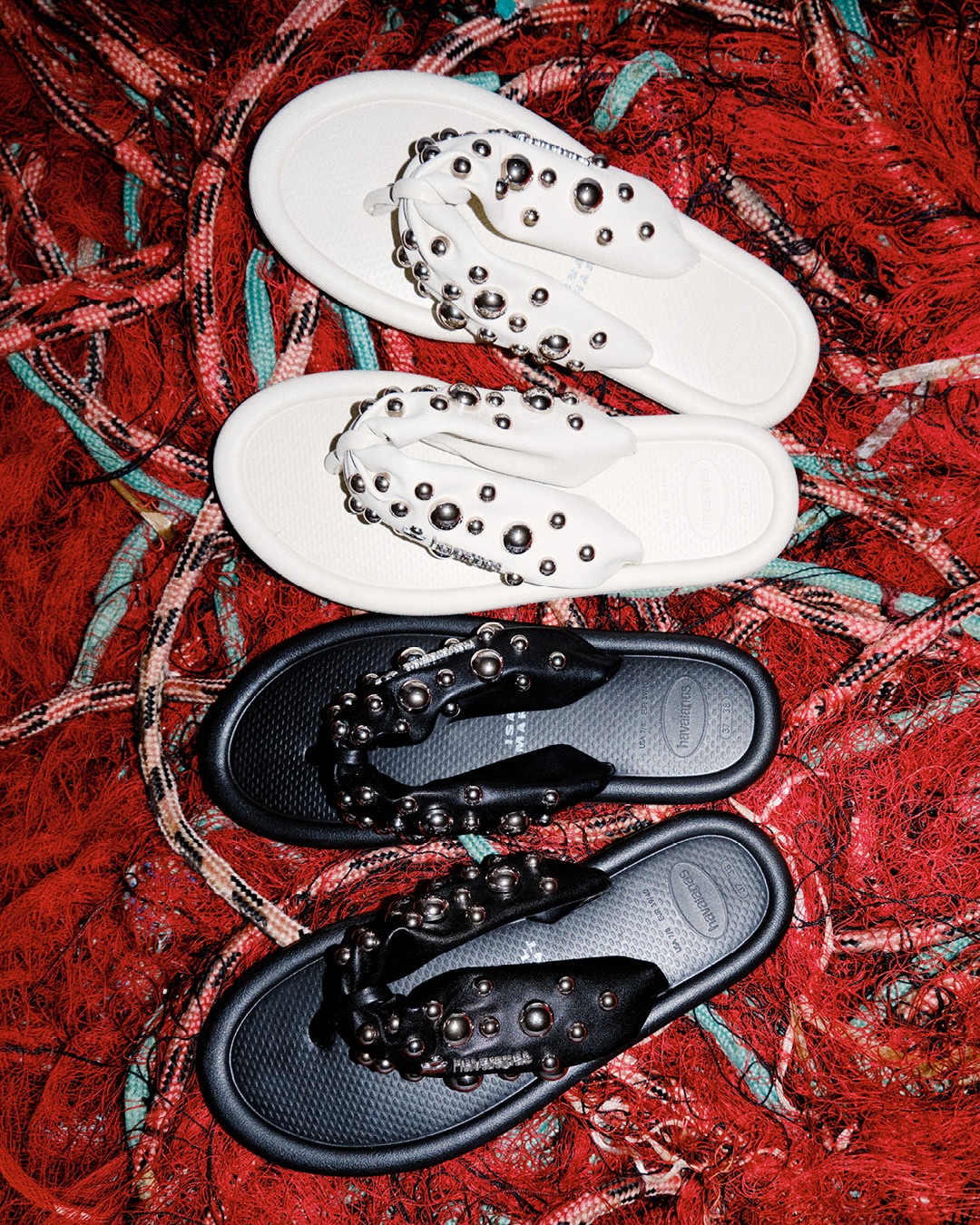 Pairs of white and black Isabel Marant x Havaianas flip-flop sandals with silver studs resting on tangled red fishing nets.