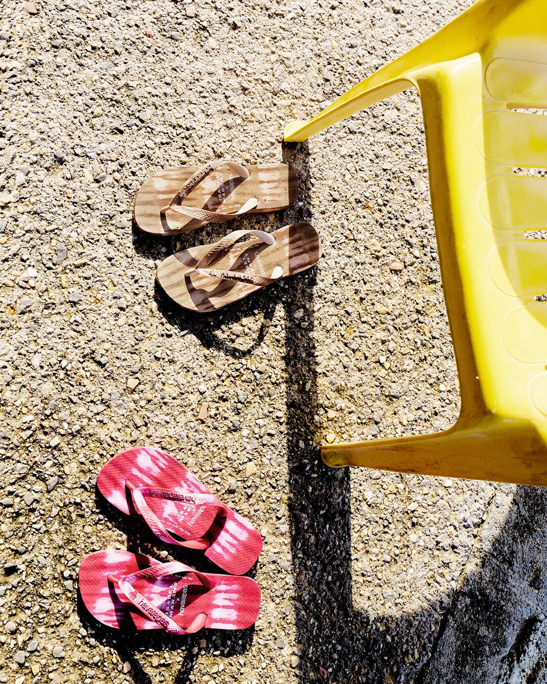 Two pairs of patterned Havaianas flip-flops rest on a concrete surface next to a bright yellow plastic chair in direct sunlight.