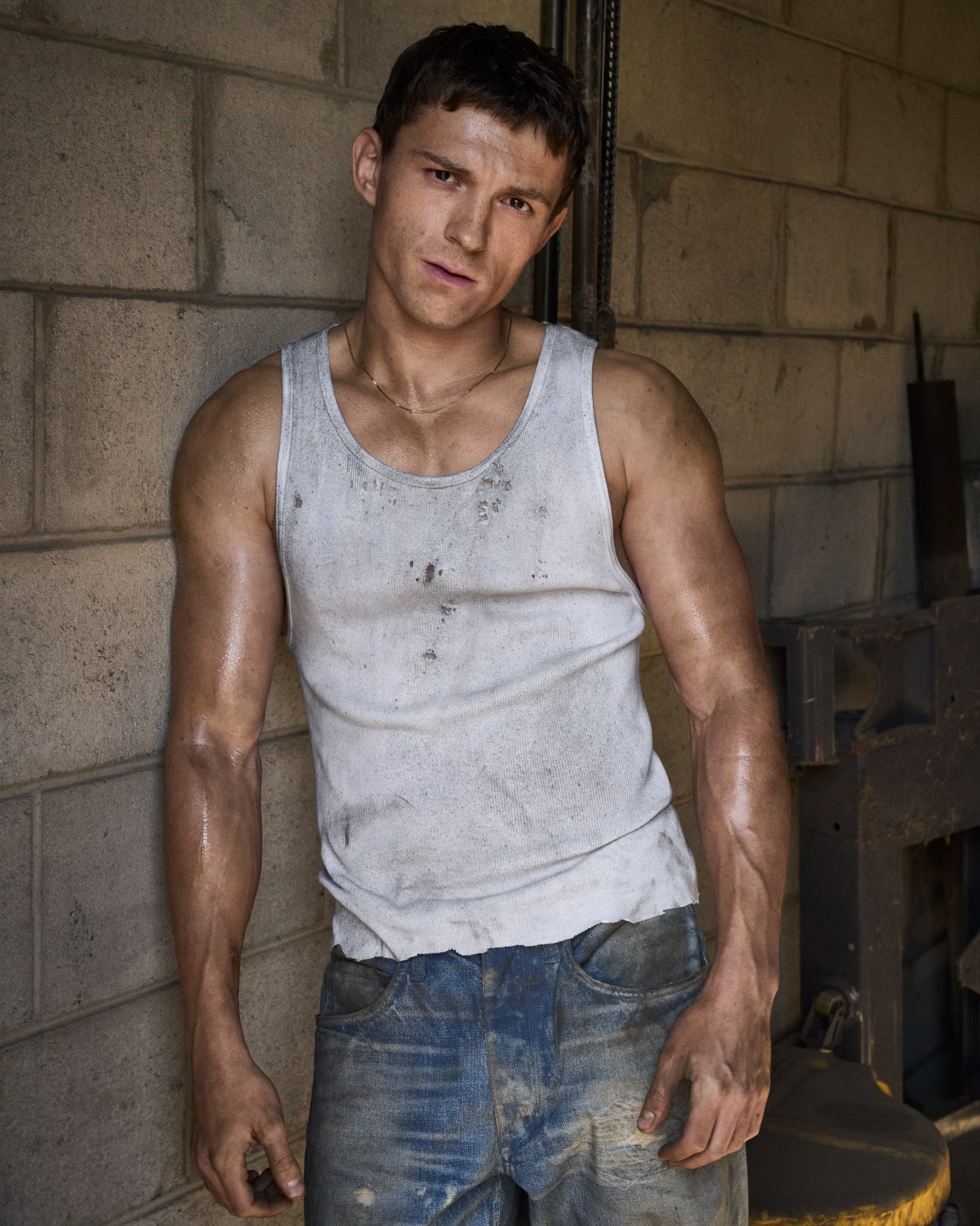 Tom Holland wearing a dirty white tank top and blue jeans leaning against a concrete cinder block wall.