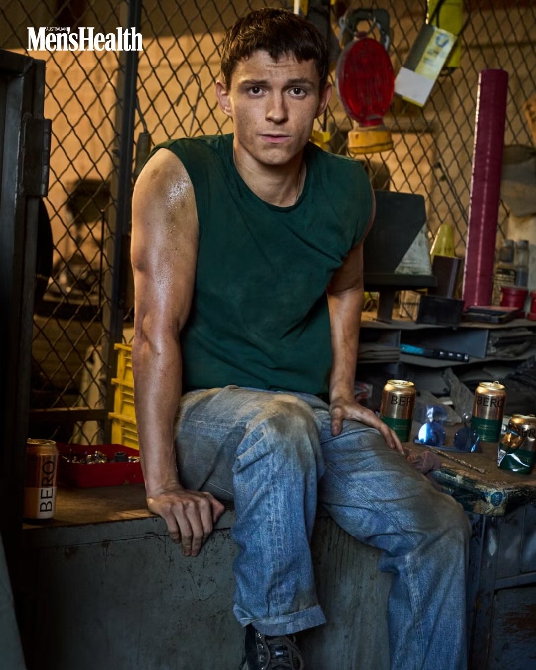 Tom Holland wearing a green sleeveless shirt and blue jeans seated on a counter in a workshop setting.