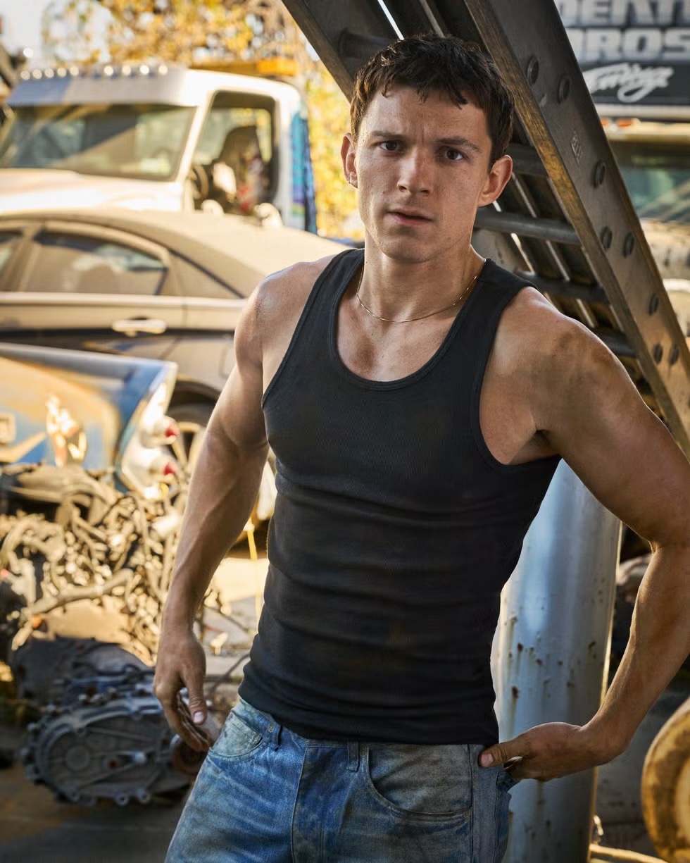 Tom Holland wearing a black tank top and blue jeans standing in a vehicle junkyard with cars and metal structures