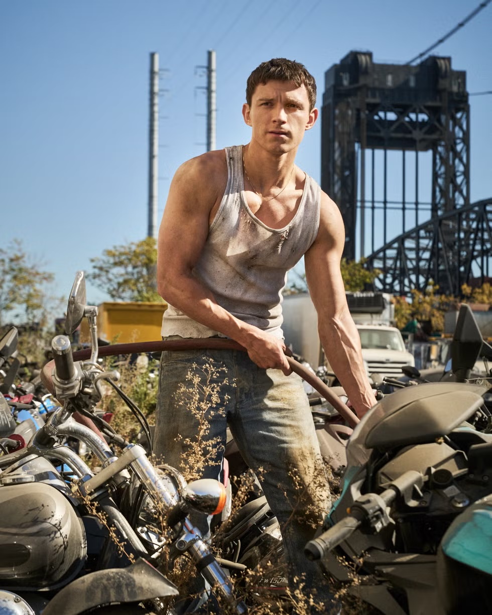 Tom Holland in a distressed white tank top and blue jeans standing among motorcycles in an industrial outdoor setting.