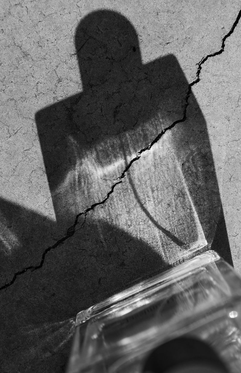 Black and white photograph of a glass perfume bottle casting a sharp bottle-shaped shadow across cracked concrete.