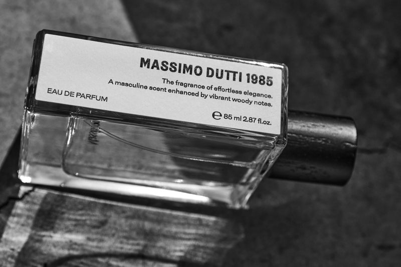 Close-up black and white photograph of a glass Massimo Dutti 1985 Eau de Parfum perfume bottle lying on a textured surface.