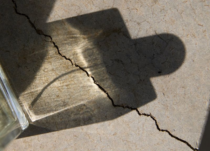 Sunlight casting the shadow and caustic light refraction of a glass perfume bottle across a cracked stone surface.