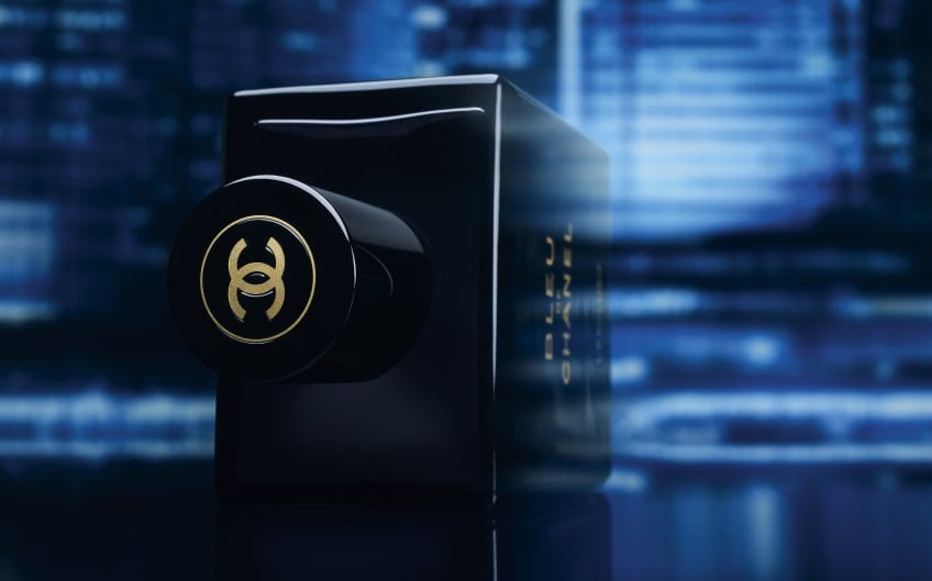 Bleu de Chanel perfume bottle with gold interlocking logo on cap against a blurred cool blue city backdrop.