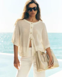 Andreea Diaconu in a cream knit cardigan, white trousers, and dark sunglasses holding a textured tote bag by the ocean.