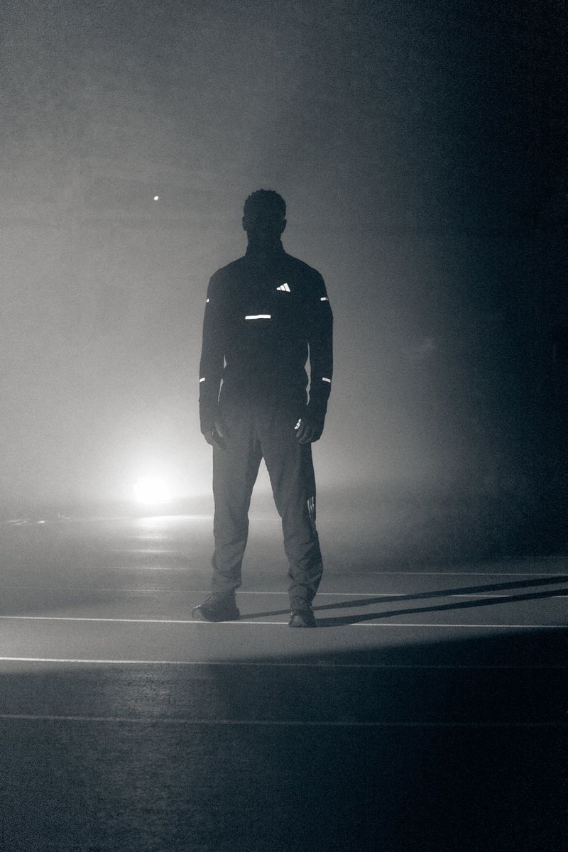 Silhouette of a man in reflective Adidas running gear standing on a dark, foggy track illuminated by strong backlight.