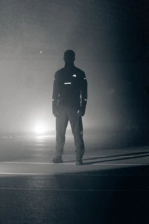 Silhouette of a man in reflective Adidas running gear standing on a dark, foggy track illuminated by strong backlight.