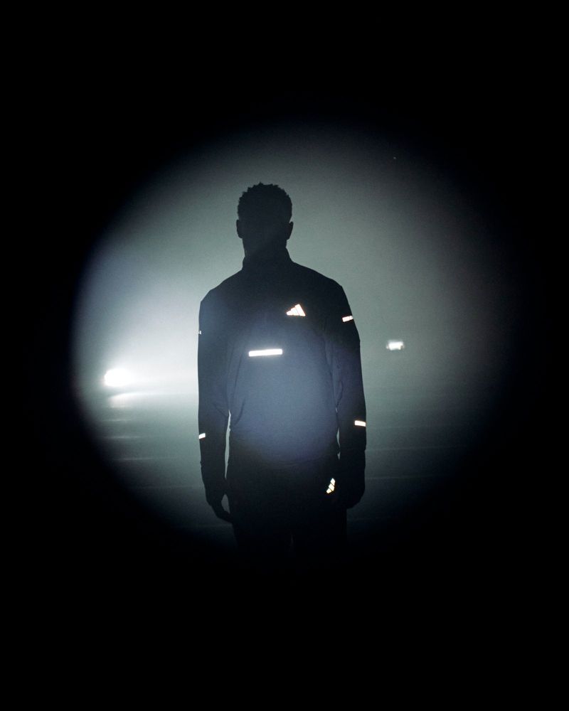 Silhouette of a person wearing a dark athletic jacket with glowing reflective markings in a circular beam of light surrounded by darkness.