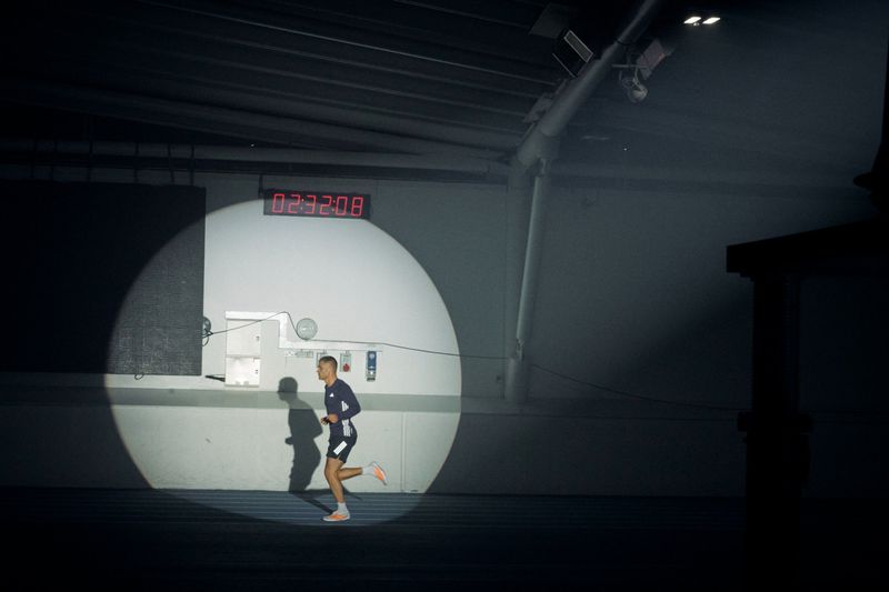 A male runner illuminated by a circular spotlight running along a dark indoor track beneath a digital timer display.