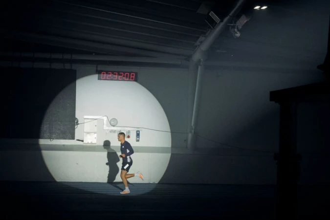 A male runner illuminated by a circular spotlight running along a dark indoor track beneath a digital timer display.
