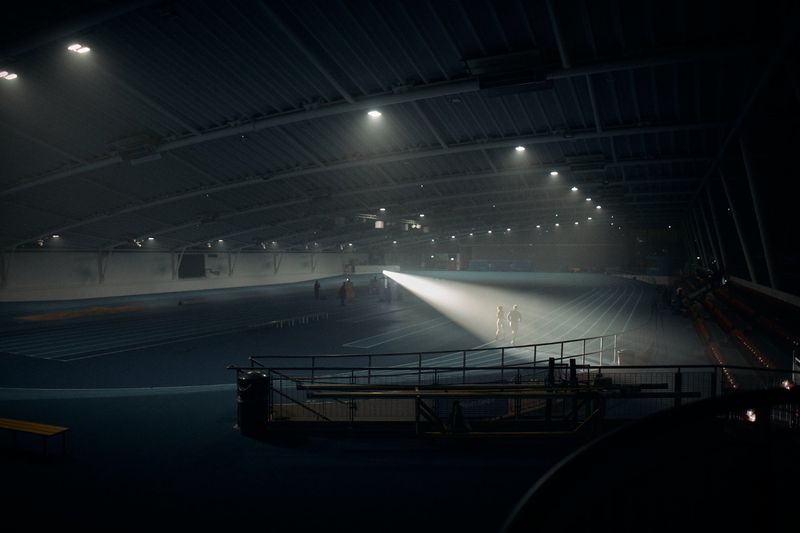 Two figures illuminated by a sharp spotlight beam on a dark indoor running track in an empty sports arena.