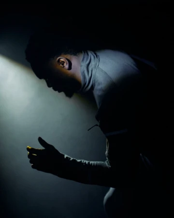 Side profile silhouette of a Black man in a high-neck grey athletic top illuminated by dramatic low-key lighting in a dark space.