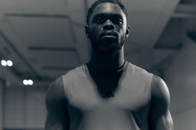 Portrait of a Black man with a beard wearing a grey sleeveless athletic top against a dark interior setting.