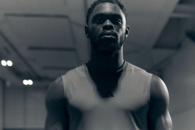Portrait of a Black man with a beard wearing a grey sleeveless athletic top against a dark interior setting.