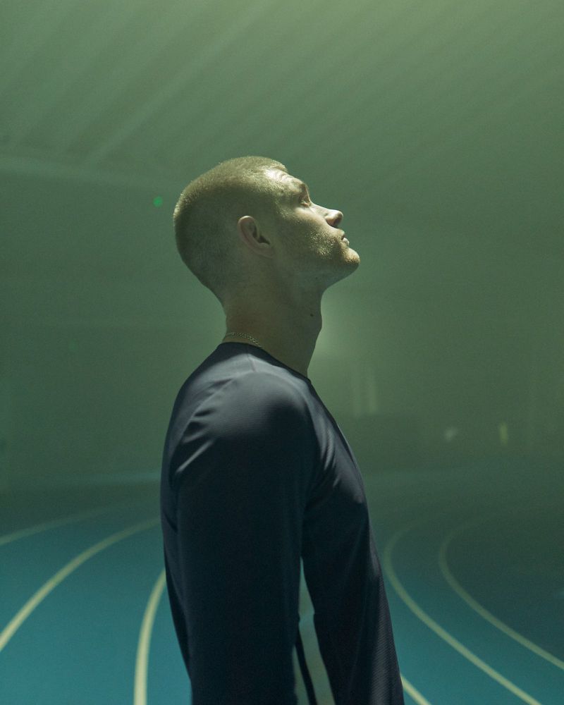 Man with a buzz cut wearing a dark long-sleeve athletic shirt looking up on a curved indoor running track in hazy green light.