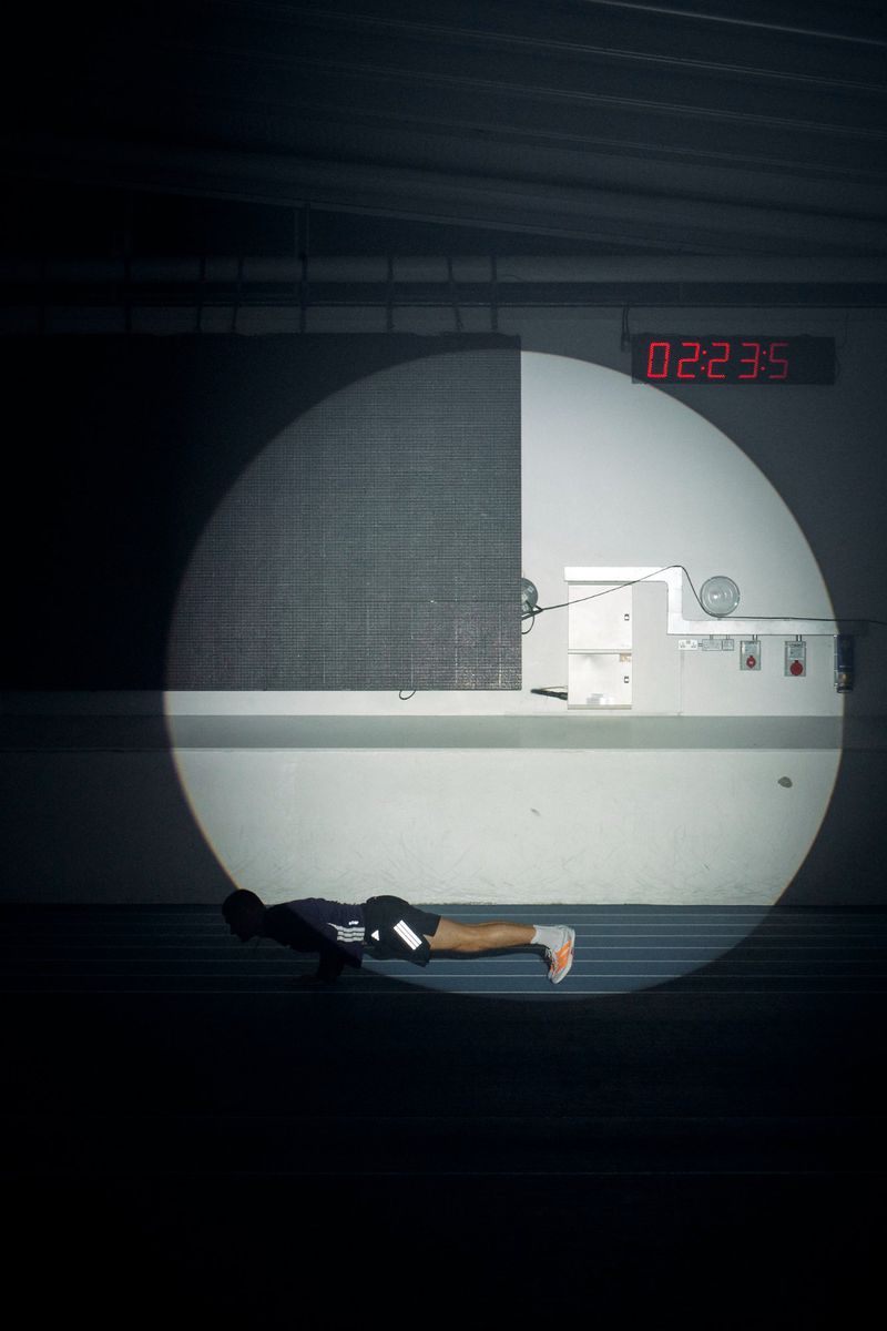 Athlete in black Adidas sportswear doing pushups inside a circular spotlight on an indoor track with a red digital clock.