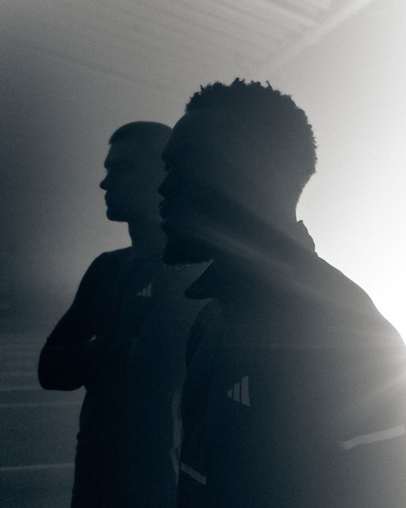 Two men in athletic wear stand together in dim lighting, showcasing Adidas branding on their clothing. The image evokes a sense of fitness.